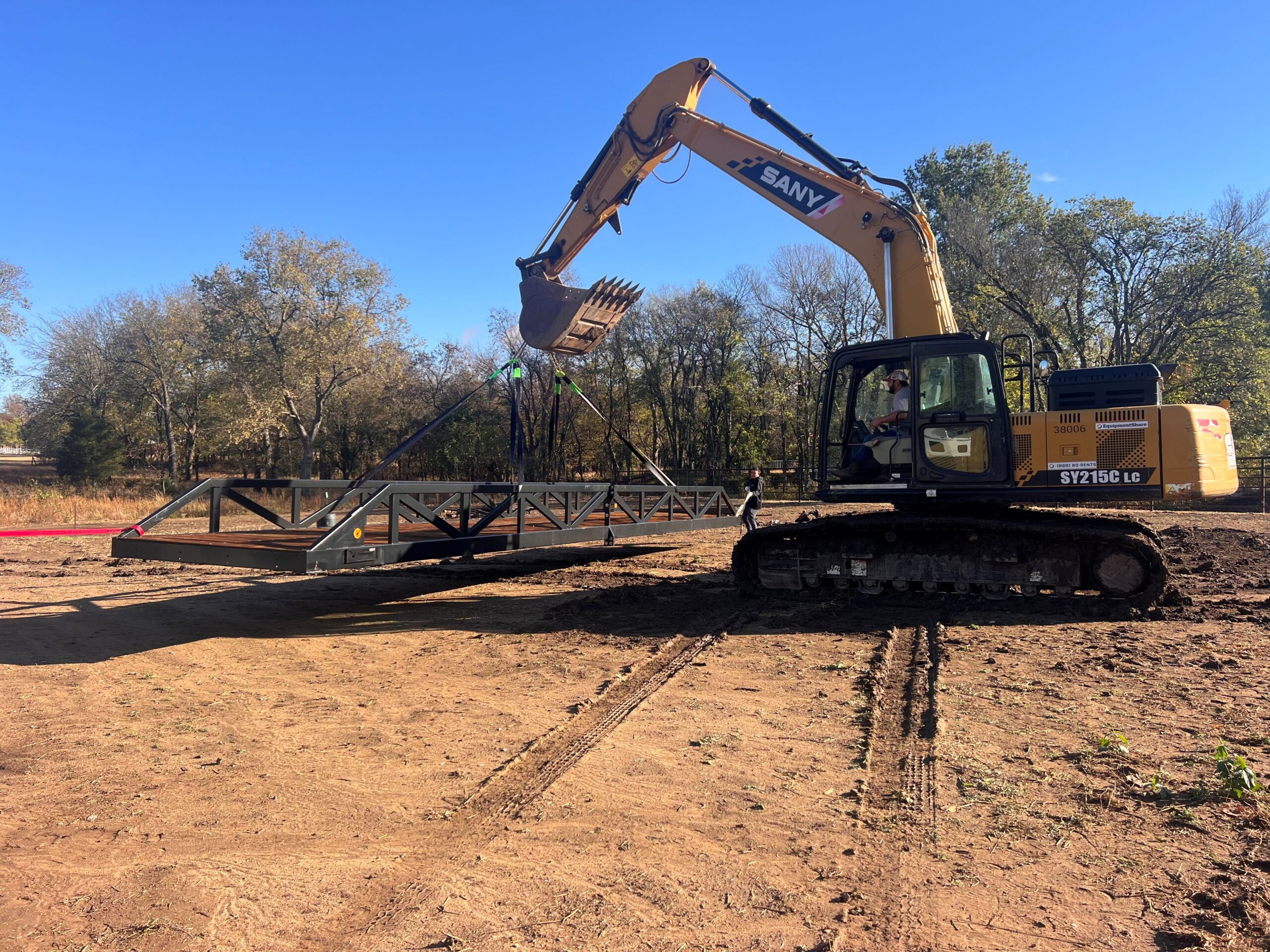 Excavator lifting and positioning a fully assembled steel truss bridge during installation on a rural site, with crew guiding the span into place.